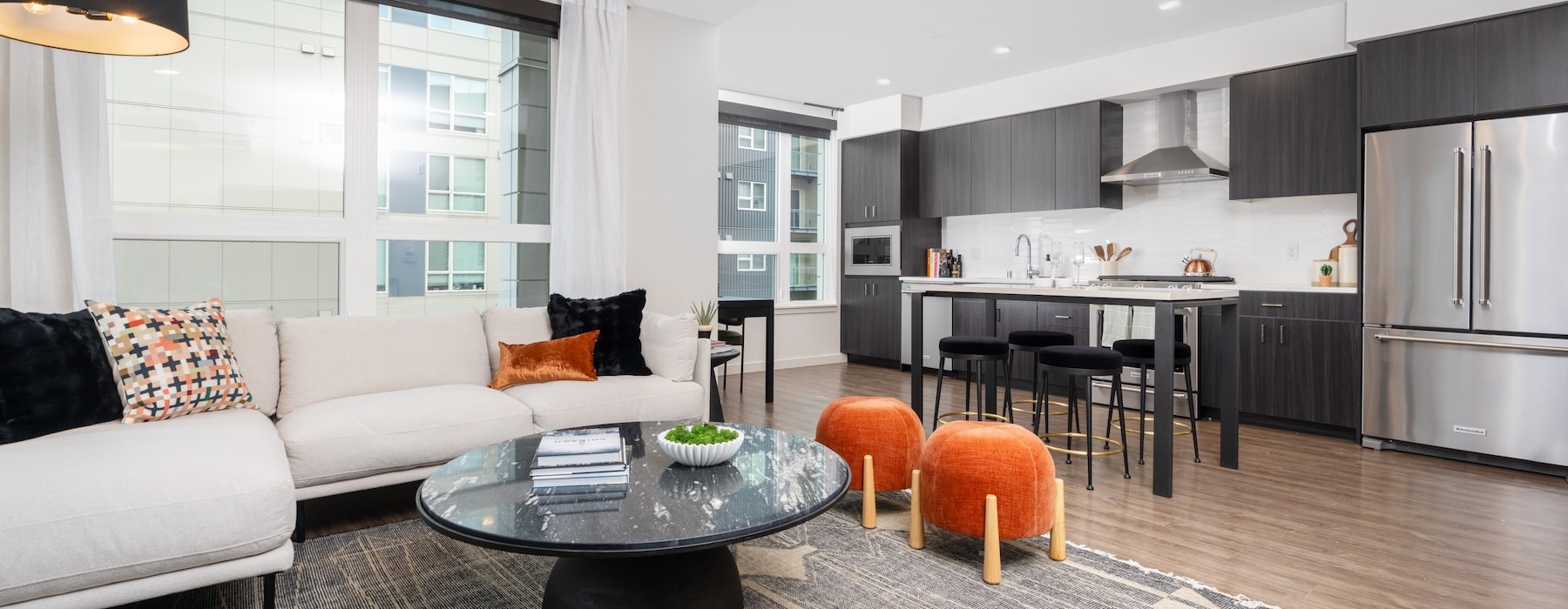 Living room with beige sectional, marble coffee table, and orange ottomans, opening into a modern kitchen with dark cabinets and a bar island.